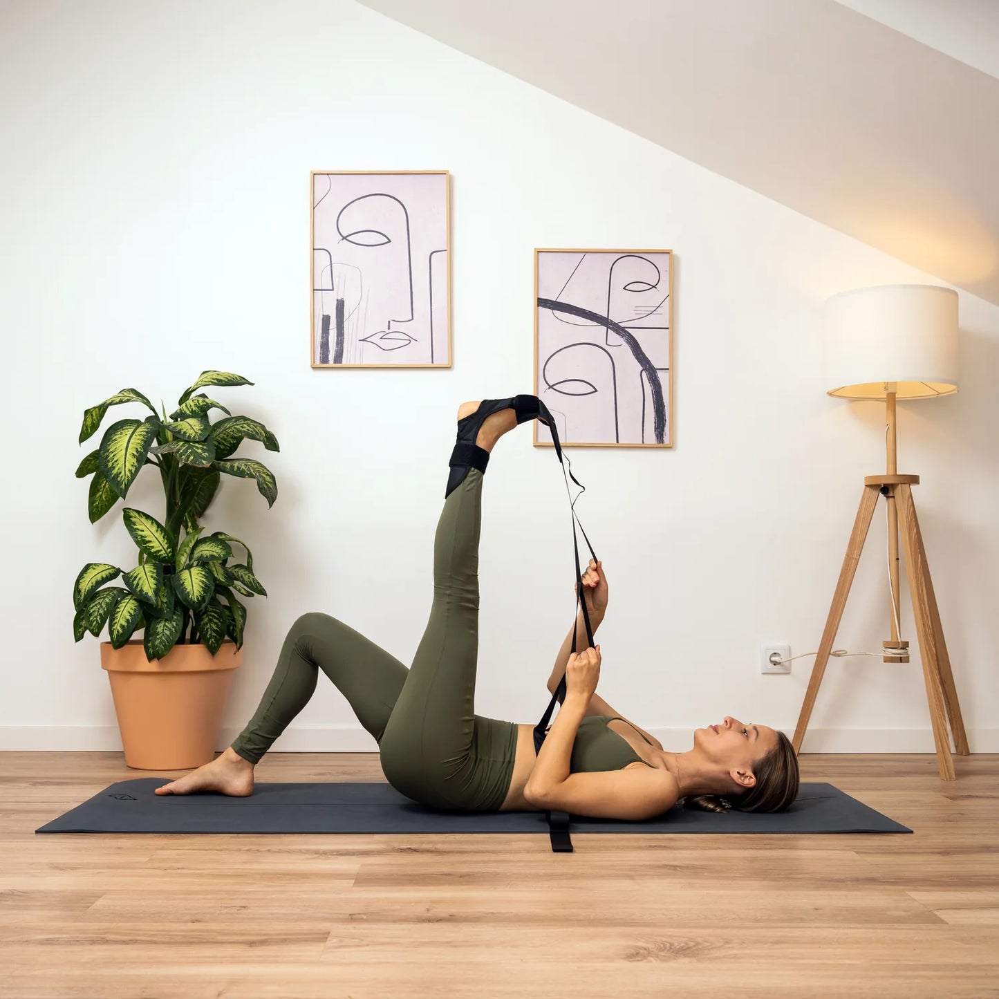 A person lying on a yoga mat using the Somabody FlexiStrap to stretch, with the strap around their left foot, while plants and a lamp are visible in the background.